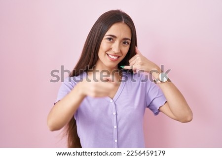 Young hispanic woman with long hair standing over pink background smiling doing talking on the telephone gesture and pointing to you. call me. 