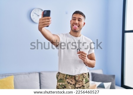 Young hispanic man wearing camouflage army uniform taking selfie at home sticking tongue out happy with funny expression. 