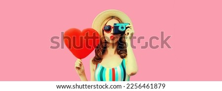 Summer portrait of young woman taking picture on film camera with big red heart shaped balloon wearing straw hat on pink background