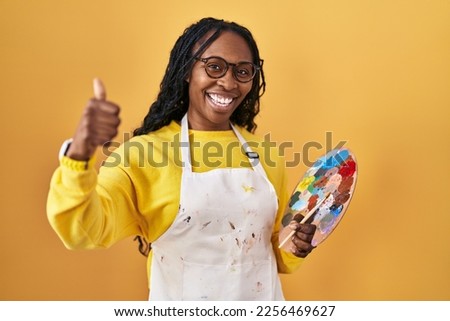 African woman holding painter palette approving doing positive gesture with hand, thumbs up smiling and happy for success. winner gesture. 