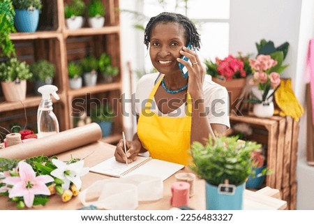 Middle age african american woman florist talking on smartphone writing on notebook at flower shop