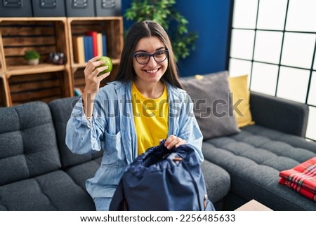 Young hispanic woman student holding apple of backpack sitting on sofa at home