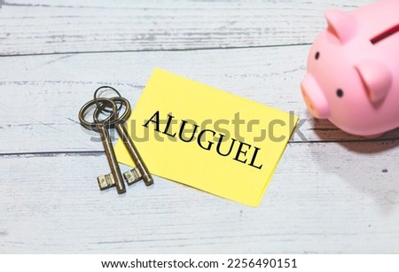 The word rent in Brazilian Portuguese written on a piece of paper on a wooden table. Keys to a house and a piggy bank in the composition.
