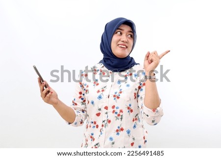 Happy asian muslim woman holding a cell phone while pointing and looking back. Isolated on white