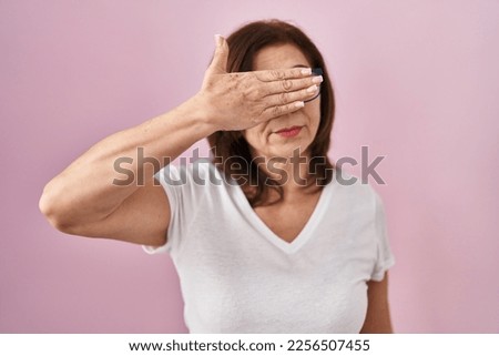 Middle age hispanic woman standing over pink background covering eyes with hand, looking serious and sad. sightless, hiding and rejection concept  Royalty-Free Stock Photo #2256507455