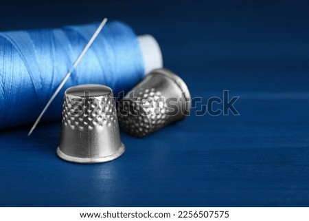 Silver thimbles, sewing thread and needle on blue wooden table, closeup. Space for text Royalty-Free Stock Photo #2256507575