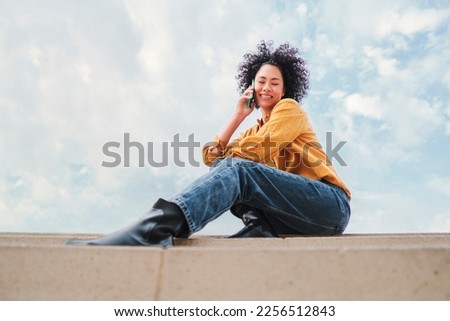 Front view of young hispanic woman with curly hair anda having a cellphone call sitting under the blue sky with clouds. Happy latin girl with yellow shirt talking by phone, at background the skyline