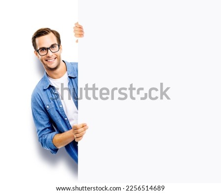 Portrait image of happy smiling casual man in eye glasses hold, peep out, stand behind mockup blank signboard, billboard, isolated over white background. White empty ad sign board banner. IT Expert Royalty-Free Stock Photo #2256514689