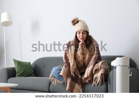 Frozen young woman with plaid warming near electric heater at home