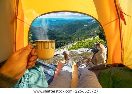 Cup of hot drink in the hand and wonderful view of mountain tops through the open entrance of the tent. The beauty of a romantic hike and camping accompanied by a dog.