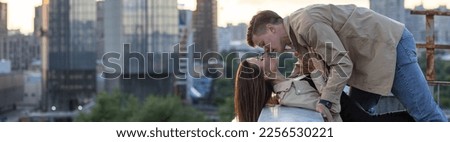 Young happy beautiful loving couple on a surprise romantic date on a rooftop on Saint Valentine's Day. Attractive man and woman, male and female hugging, kissing. Sunset, skyscrapers urban view banner