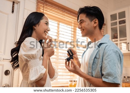 Asian romantic man making surprise proposal of marriage to girlfriend. Attractive young male and proposing to beautiful happy woman, with wedding ring enjoying surprise engagement in kitchen at home. Royalty-Free Stock Photo #2256535305