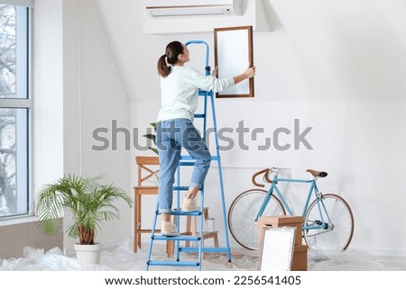 Young woman on ladder hanging frame onto light wall at home