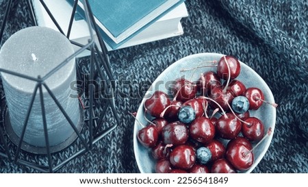 Bowl full of ripe red cherries candlestick and books on a dark blue knitted blanket. View from above.