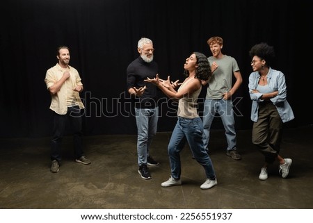 multiracial woman gesturing while rehearsing near bearded art director and smiling interracial actors Royalty-Free Stock Photo #2256551937
