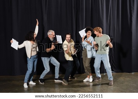 overjoyed multiracial theater troupe holding clipboards with screenplays and showing success gesture. Translation of tattoo: om, shanti, peace Royalty-Free Stock Photo #2256552111
