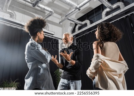 positive and emotional acting skills teacher gesturing near young interracial women in theater. Translation of tattoo: om, shanti, peace Royalty-Free Stock Photo #2256556395