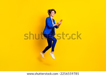 Full length photo of good mood woman wear blue jacket typing samsung apple device jumping high isolated yellow color background