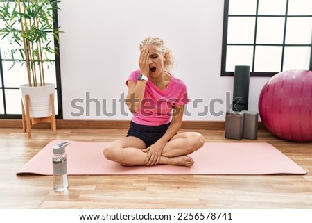 Middle age blonde woman sitting on yoga mat yawning tired covering half face, eye and mouth with hand. face hurts in pain.  Royalty-Free Stock Photo #2256578741