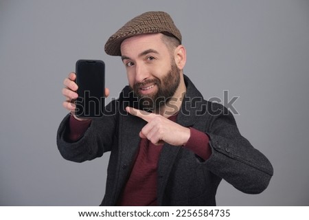 Stylish young man in jacket and cap pointing his cell phone screen to the camera extending his arm forward in a studio against a gray background.