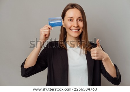Young cheerful woman is holding thumb up and recommending a blue credit card over grey background.