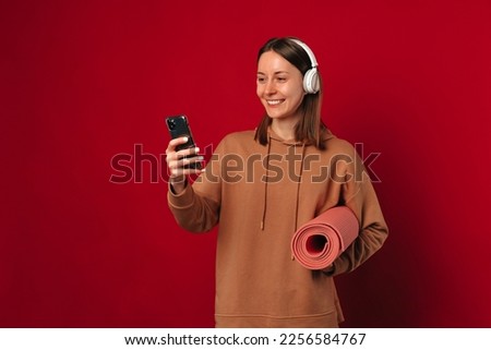  Ecstatic young woman wearing headphones is holding her fitness mat and phone over red background.