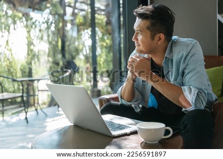 Asian handsome man sitting happily looking out of the window in a cafe. Concept freelance and financial freedom. Royalty-Free Stock Photo #2256591897