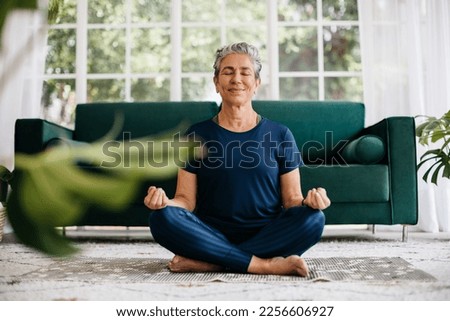 Senior woman meditating in lotus position at home, sitting on the floor in fitness clothing. Mature woman doing a breathing workout to achieve relaxation, peace and mindfulness in a healthy lifestyle. Royalty-Free Stock Photo #2256606927