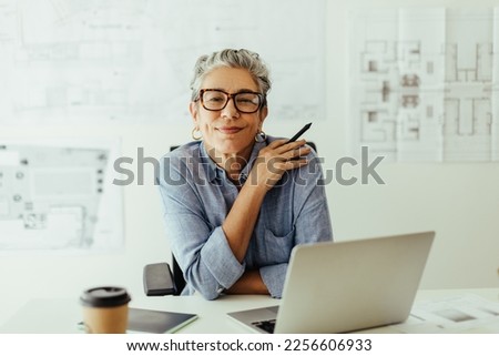 Mature female designer looks at the camera in her office, confident in her expertise and style of creativity. Senior design professional using a laptop and a graphics tablet to put her skills to work. Royalty-Free Stock Photo #2256606933