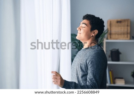 Portrait of positive hispanic teenager boy breathing deep indoors beside the window