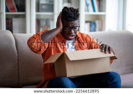 Disappointed unhappy african american young guy customer sitting on couch at home, holding paper box delivery on his lap, opening parcel and gestruring, dissatisfied with order from webstore Royalty-Free Stock Photo #2256612895