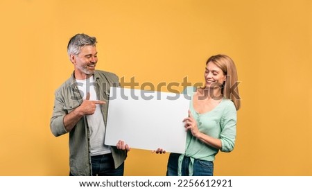 Place for your ad. Middle aged couple holding empty placard board, man pointing on it, yellow studio background. Spouses standing with white paper, free space Royalty-Free Stock Photo #2256612921