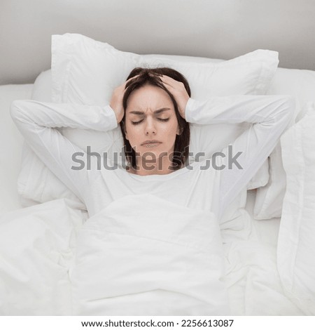 Top view of unhappy young brunette woman lying in bed at home, touching head with both hands, tired lady wake up with headache in the morning, suffering from chronic migraine, copy space Royalty-Free Stock Photo #2256613087