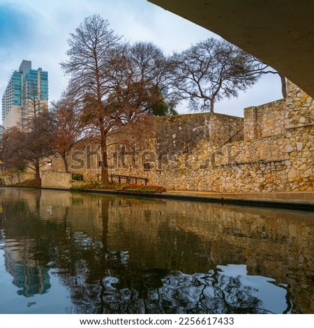 Tranquil San Antonio River Walk footpath, stonewall landscape feature, and boardwalk water reflections with city skyline and barren trees in winter along the canal in Texas, off-season cityscape
