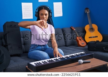 African american woman with braids playing piano keyboard at music studio with angry face, negative sign showing dislike with thumbs down, rejection concept 