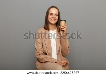 Image of attractive joyful cheerful woman wearing beige jacket posing isolated over gray background, sitting with take away coffee in hands, enjoying hot beverage.