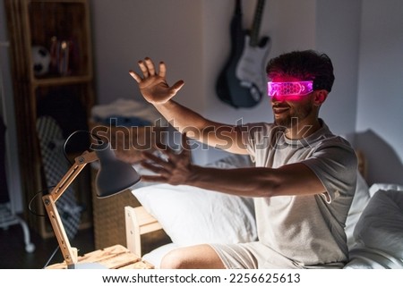 Young arab man using augmented reality glasses sitting on bed at bedroom