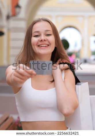 Vertical photo of Happy blurred young woman is showing, holding credit bank card in hand and smiling during shopping with shopping bags in mall