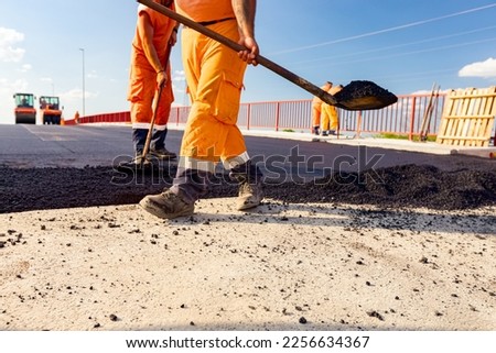 Few workers are using shovels to level, set up layer of fresh tarmac to right measures, pouring hot asphalt. Royalty-Free Stock Photo #2256634367