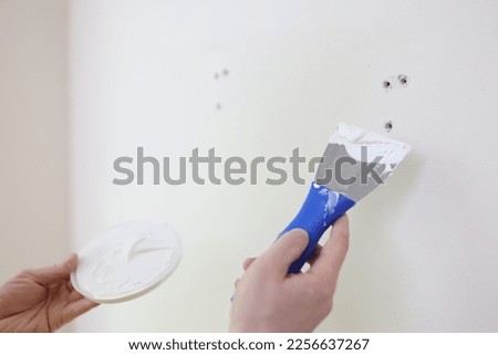 Hands with a spatula and putty against the background of a white wall, a close-up. Scatter holes on the wall Royalty-Free Stock Photo #2256637267