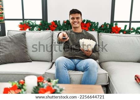 Young hispanic man watching tv sitting on sofa by christmas decor at home