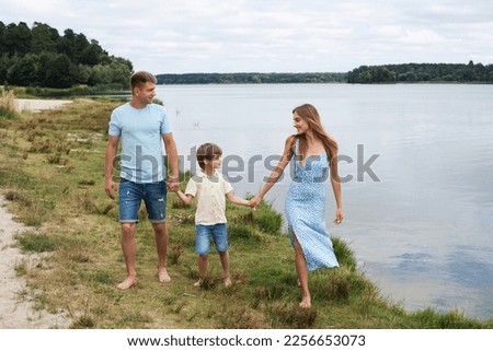 Portrait of a happy family in nature, walking on the banks of a picturesque river. Mom is dressed in a summer dress, dad and son are in shorts and t-shirts.