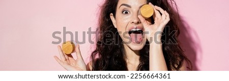 Shocked young woman holding jars with cosmetic creams on pink background, banner Royalty-Free Stock Photo #2256664361