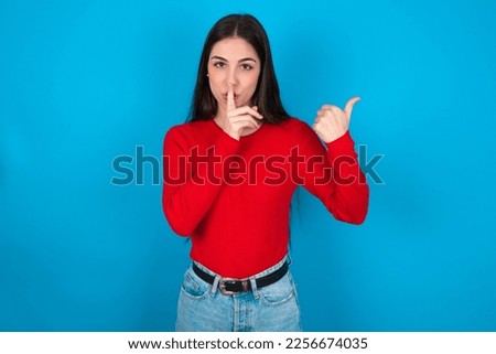 young caucasian brunette girl wearing red T-shirt against blue wall asking to be quiet with finger on lips pointing with hand to the side. Silence and secret concept.