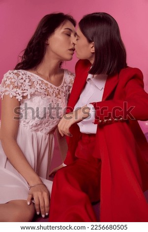  Valentine's day concept. Studio portrait of two young lesbian girls enjoying intimate moment together on a pink background. One girl is in a white dress, the second woman is in a red suit.           