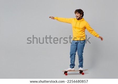 Full body young young happy fun cool Indian man 20s he wearing casual yellow hoody riding skateboard pennyboard look camera isolated on plain grey background studio portrait. People lifestyle portrait