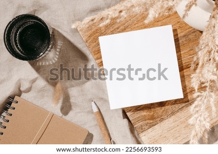 Blank paper sheet card mockup on wooden desk, vase with dried lagurus grass, notepad and glass of water with sunlight shadows on beige linen cloth. Minimal aesthetic brand template, home office desk