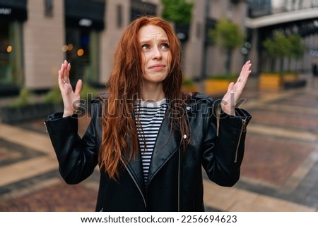 Portrait of frustrated red-haired young woman touching wet hair after autumn rain standing on beautiful city street. Front view of upset female untangling hair after being caught in rain. Royalty-Free Stock Photo #2256694623