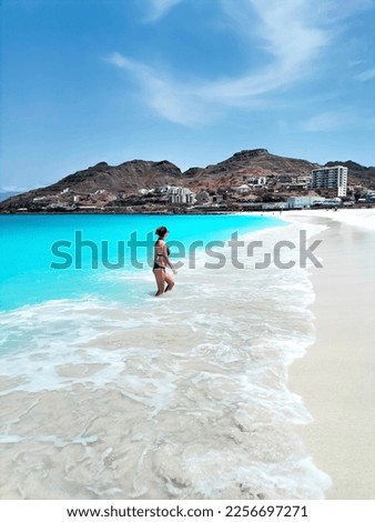 Young woman in swimsuit coming out of the turquoise ocean in a foaming wave toward the white sandy beach against the backdrop of mountains in Mindelo, Sao Vicente island, Cape Verde.