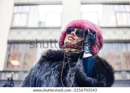 Fashionable pretty woman in sunglasses wearing natural fur coat and pink fur hat at city building background, looking up away. Stylish young lady posing outdoor. Fashion style concept. Copy text space